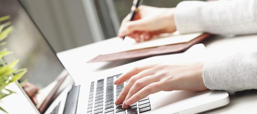 A woman types on a laptop computer with her left hand as the takes notes in a notebook using a pen with her right hand.